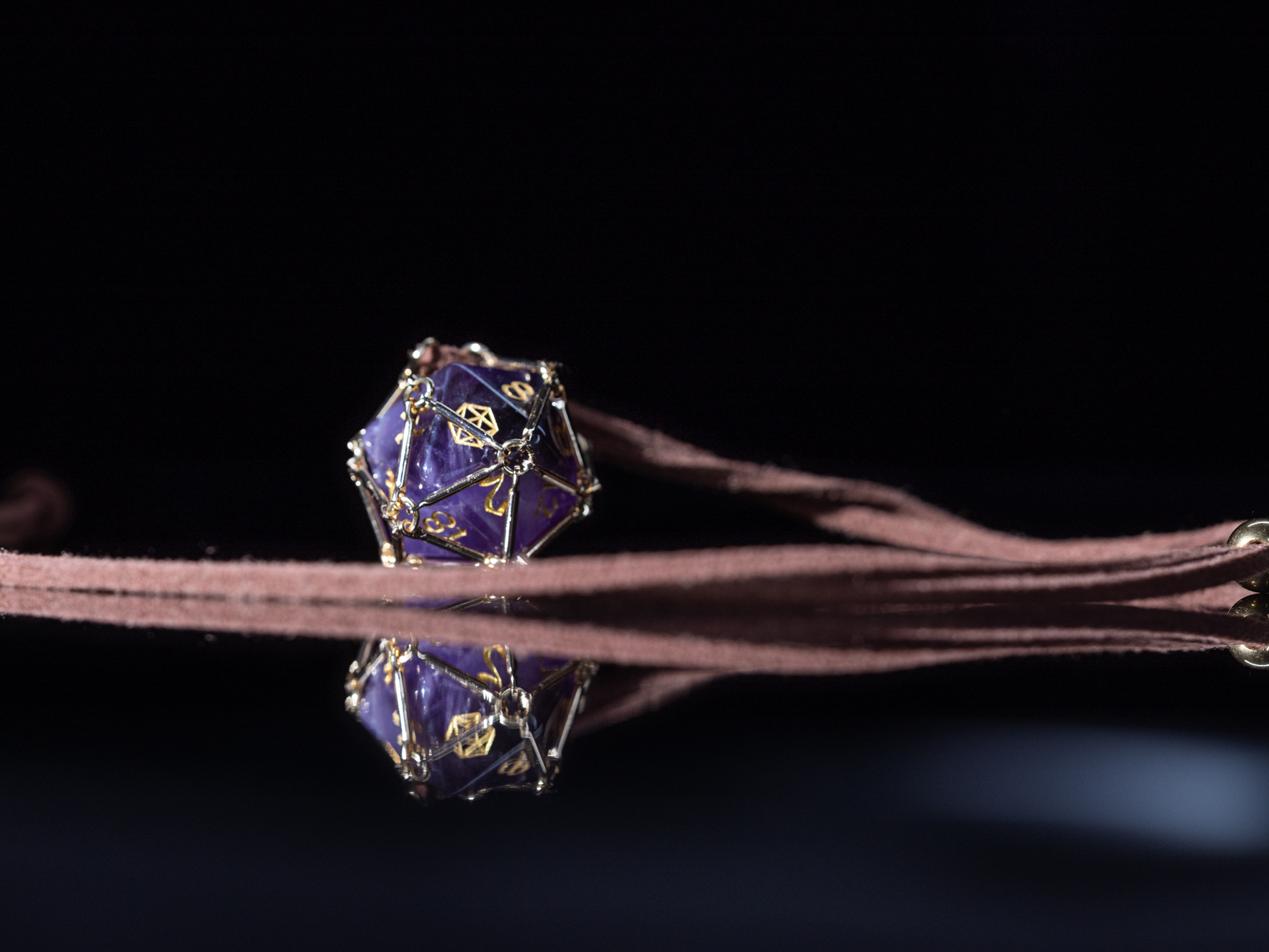 Angled macro shot of “Saga – Arcane Focus”: purple Amethyst D20 inside a golden dice cage on a brown PU leather necklace.