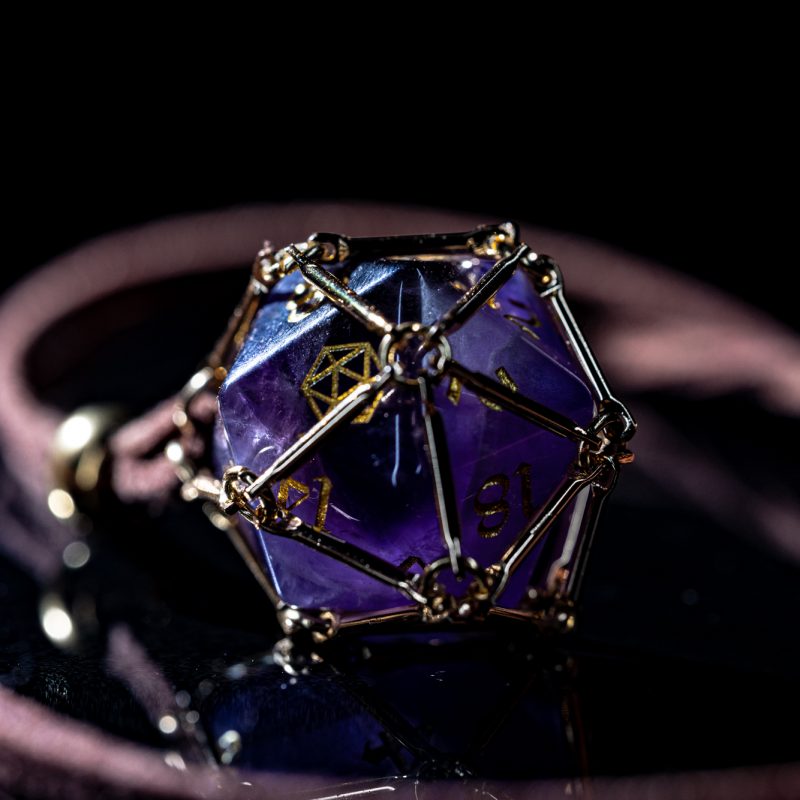 Angled macro shot of “Saga – Arcane Focus”: purple Amethyst D20 inside a golden dice cage on a brown PU leather necklace.